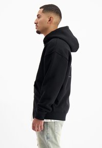 Zwarte hoodie met een losse pasvorm, ribbelboorden en een geribde onderkant. Kenmerkt zich door een kangoeroe-zak en een eenvoudig ontwerp, gedragen over lichte denim jeans.