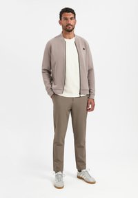 Lichtgrijze bomberjack met ritssluiting, gecombineerd met een textuurwit sweater, beige broek en grijze sneakers met witte accenten.