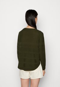 Olivgrüner, strukturierter Pullover mit Rundhalsausschnitt, langen Ärmeln und leicht abgerundetem Saum, kombiniert mit hellen Shorts.