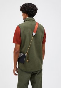 Green fleece vest with a high collar, red T-shirt underneath, and a black pouch secured by an orange cord on the back.