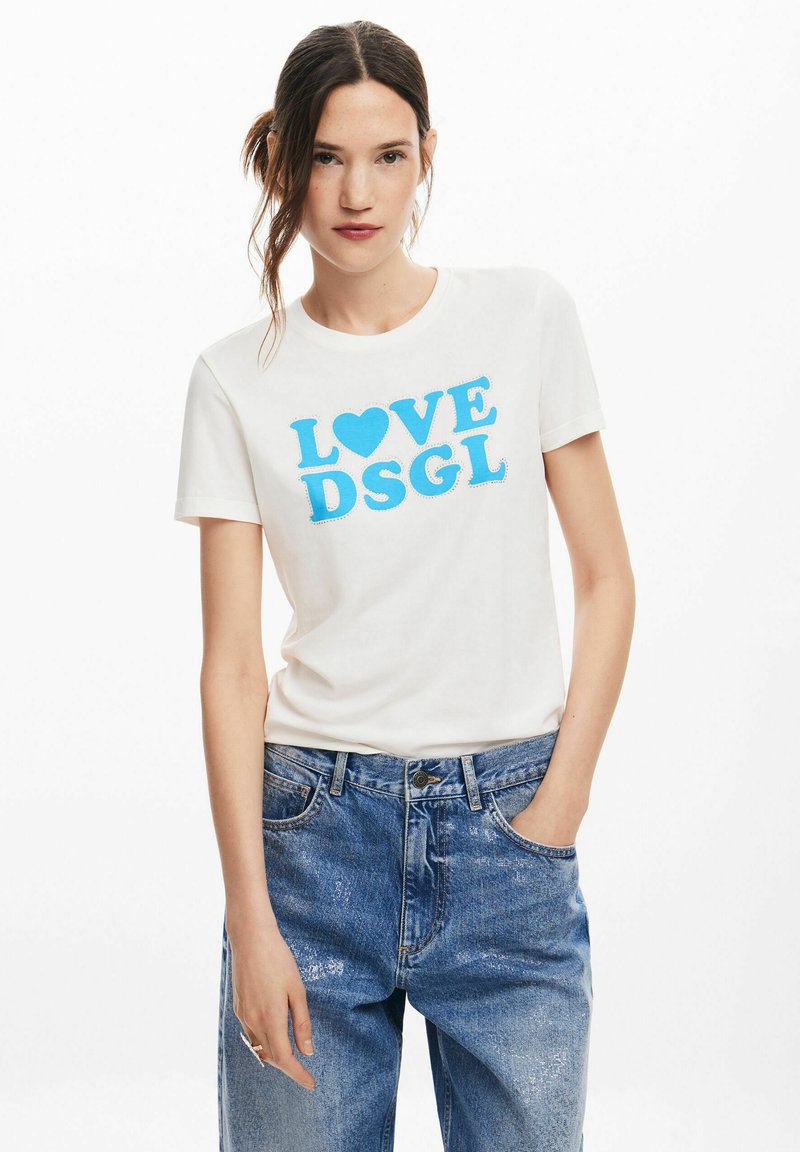 T-shirt bianco in cotone con il testo blu "LOVE" e un simbolo a cuore. Maniche corte, scollo rotondo, vestibilità rilassata. Indossato con jeans blu.