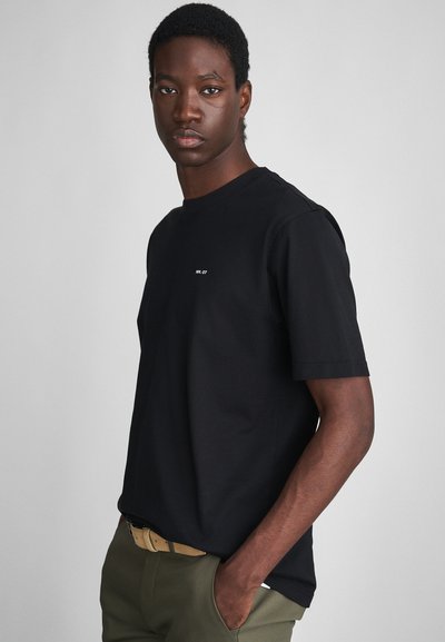 Jeune homme portant un t-shirt noir avec le texte "NN.07", un pantalon vert olive et une ceinture beige, se tenant avec une main dans la poche.