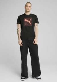 T-shirt in cotone nero con un design grafico rosso del logo Puma all'interno di una griglia. Abbinato a jeans neri a gamba larga e sneakers nere.