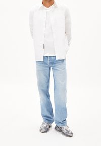 Camisa blanca de botones sobre una camiseta blanca, combinada con jeans descoloridos de color azul claro y zapatillas grises con acentos negros.