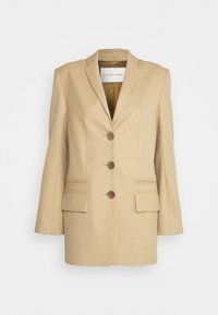 Blazer beige sur mesure avec revers à crans, trois boutons marron, deux poches frontales avec couture horizontale et une texture lisse.