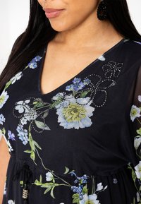 Schwarzes Blumenkleid mit V-Ausschnitt, leuchtend blauen und grünen Blumenmustern sowie dekorativen Perlenakzenten auf dem Stoff.