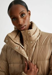 Giubbotto imbottito beige con colletto alto, chiusura con zip e bottoni a pressione. Texture liscia con dettagli trapuntati.