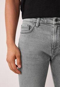 Szürke farmernadrág, testhez simuló szabással, részletes varrással, öt zsebbel és övbújtatóval, egy kéz tartja méretarányként.