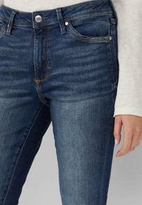 Jean en denim bleu foncé avec une taille haute, coupe slim, présentant des coutures dorées, des poches latérales et un fermoir à bouton visible à la taille.