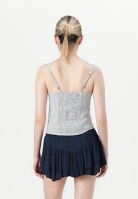 Mulher com cabelo loiro apanhado em um coque, vestindo uma blusa sem mangas cinza às riscas e uma saia em camadas azul marinho, vista de costas contra um fundo branco.