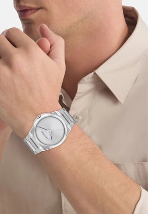 Montre analogique argentée avec un cadran rond et minimaliste, des aiguilles bleues et un bracelet en acier inoxydable. Portée au poignet sur un fond de chemise neutre.