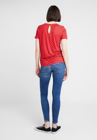Maglietta rossa a maniche corte con dettaglio a chiave sul retro, caratterizzata da un motivo a pois bianchi ripetuto. Abbinata a jeans skinny blu e sneakers nere.