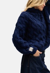 Granatowy sweter dziany z wysokim kołnierzem, z teksturowanymi wzorami warkoczy i ściągaczem na dole. Zawiera firmową metkę na rękawie.