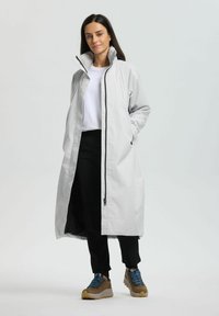 Mujer de pie con las manos en los bolsillos, vestida con un abrigo largo de color gris claro, camisa blanca, pantalones negros y botas de senderismo marrones con cordones azules.