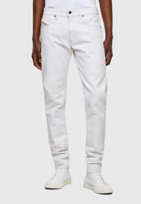 Man wearing slim-fit white jeans and white sneakers, standing against a plain light gray background.