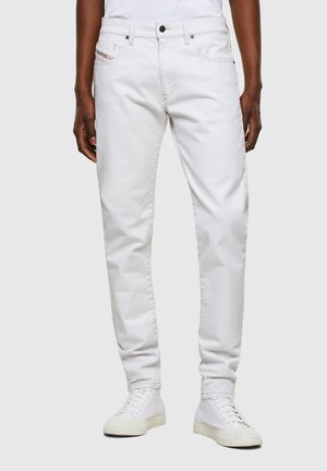 Hombre con jeans ajustados blancos y zapatillas blancas, de pie contra un fondo gris claro liso.