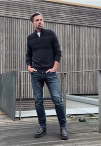 Černý svetr na zip s texturovanými horizontálními pruhy, tmavě modré džíny a černé kotníkové boty. Venkovní prostředí s dřevěným pozadím.