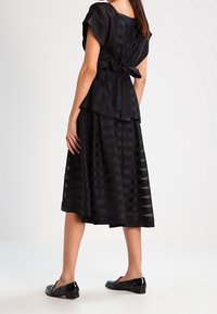 Černé šaty s přiléhavým živůtkem a rozšířenou sukní, s texturovaným pruhovaným vzorem. Vázání v pase a krátké rukávy. Lesklé černé baleríny.