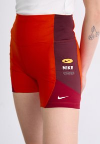 Nike aukštos juosmens šortai ryškiai raudonos ir tamsiai raudonos spalvos, su balta logo ir ženklais šone, pagaminti iš tamprių medžiagų.