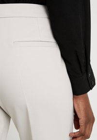 Valkoiset muotopuvun housut, joissa on sileä pinta ja takatasku. Musta pitkähihaista paita napitettavilla hihansuilla.