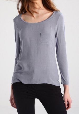 Vrouw draagt een lichtpaars shirt met lange mouwen en een klein borstzakje, gecombineerd met een zwarte broek, staand tegen een effen achtergrond.