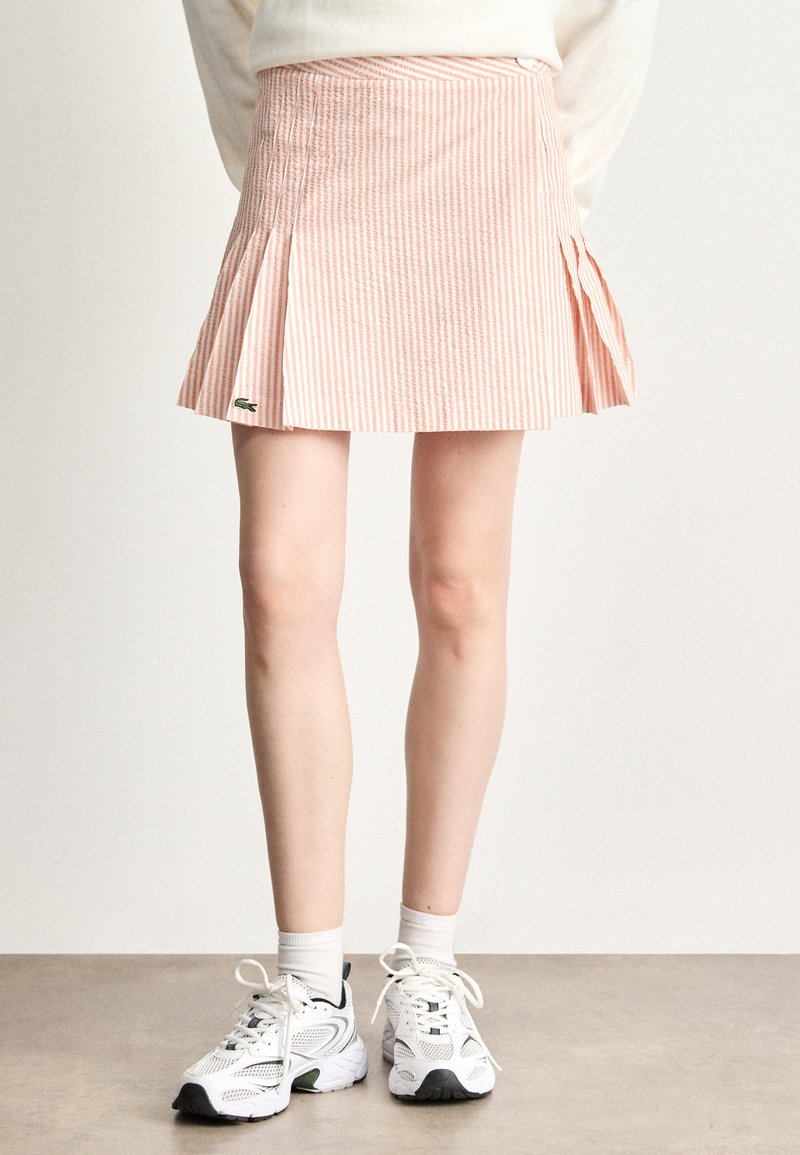Roze en wit gestreepte geplooide rok met een Lacoste-logo, gemaakt van een gestructureerde stof. G gedragen met witte sportschoenen en lichte sokken.