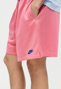 Άτομο που φοράει ροζ υφασμάτινα σορτς Nike με υφή, με τα χέρια στις τσέπες, εμφανίζεται από τους μηρούς μέχρι τη μέση σε λευκό φόντο.