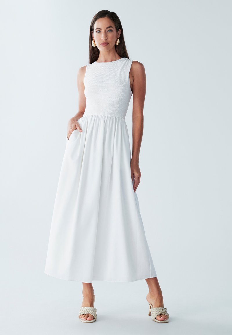 Robe maxi blanche avec un corsage ajusté et un smocké élastique, une jupe fluide, des poches latérales, stylée avec des sandales à talons tressées et des boucles d'oreilles en or.