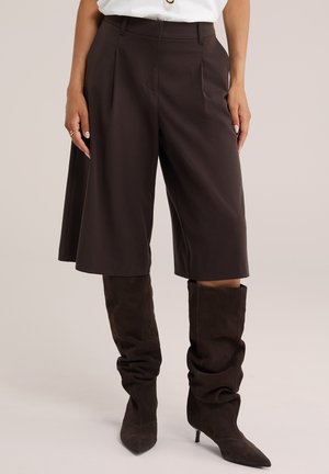 Donna che indossa pantaloncini marrone scuro a gamba larga fino al ginocchio e stivali alti con tacco e dalla vestibilità scivolata abbinati, su sfondo uniforme.