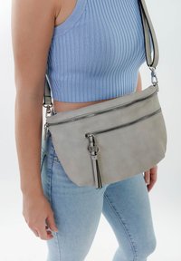 Borsa a tracolla in pelle grigia con due scomparti con zip, un accentuazione con nappina e una tracolla regolabile, abbinata a una maglietta blu a costine e jeans.