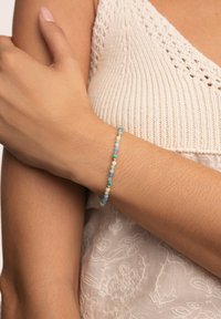 Bracciale di perline composto da una miscela di perline opache e traslucide in tonalità di blu, verde e bianco, con dettagli dorati, indossato su un polso.