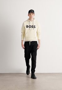 Jongeman die loopt, gekleed in een crèmekleurige hoodie met de tekst "BOSS", zwarte joggers, zwarte sneakers en een zwarte pet met witte letters.