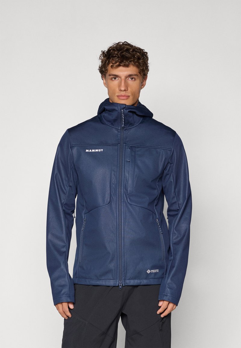 Marineblå softshell-jakke med glidelås foran, to brystlommer og hette. Har strukturert stoff og en tettsittende design.