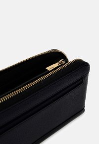 Zwarte leren portemonnee met een gestructureerd oppervlak, voorzien van een gouden ritssluiting en een externe ritstas. Compacte rechthoekige vorm met een strak ontwerp.