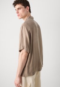 Beige rövidujjú póló, gallérral, sima textúrával, enyhén megnyújtott hátsó szegéllyel és diszkrét fényű felülettel. Világos színű nadrággal viselve.