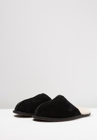 Sorte skinneslippers med en buet form, myk saueskinnforing og en teksturert såle. Har diskret merkevare på utsiden.