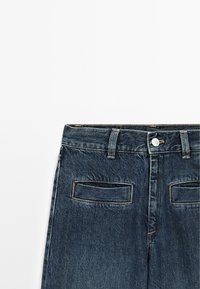 Jeans di denim blu scuro con una vita standard, bottone metallico argentato e due tasche posteriori. Tessuto testurizzato con riflessi di cuciture chiare.
