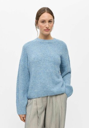 Femme aux cheveux bruns portant un pull duveteux bleu clair et un pantalon plissé gris clair, debout devant un fond blanc uni.