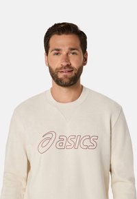 Sweatshirt beige clair avec un col rond, arborant un logo "asics" rouge. Le tissu semble doux avec une texture lisse.