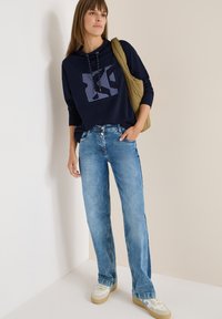 Sudadera con capucha de color marino con estampado gráfico, jeans rectos de color azul claro y zapatillas beige. Modelo sosteniendo un bolso verde oliva.