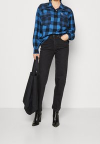 Blue and black checked flannel shirt with two chest pockets, paired with black high-waisted jeans and black heeled ankle boots, holding a black tote bag.
