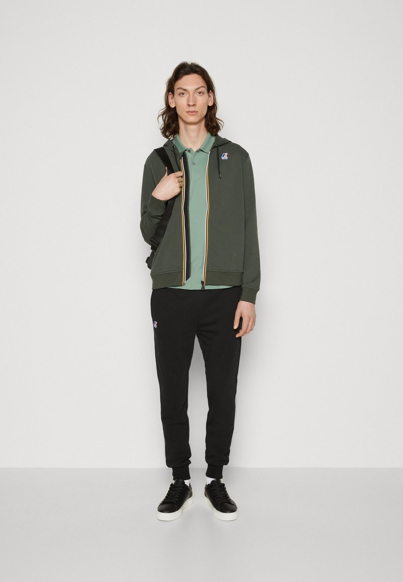Giacca verde scuro con zip e strisce multicolori, polo verde chiaro sotto, pantaloni sportivi neri e sneakers nere. Accenti di logo semplici.