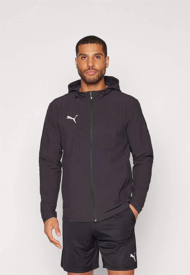 Jachetă zip-up neagră cu un model texturat, dotată cu glugă, mâneci lungi și un logo alb Puma pe piept. Purtați-o cu șorturi negre.
