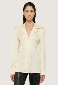 Blusa de seda crema con cuello muesca, botones frontales y mangas largas. Presenta un diseño ligeramente ajustado y una costura de contraste sutil.