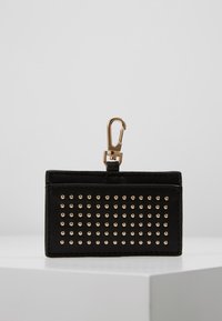 Monedero llavero rectangular negro con tachuelas doradas en filas y clip dorado en la parte superior, colocado sobre una superficie blanca frente a un fondo gris.