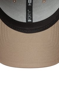 Casquette beige avec visière noire, fabriquée en tissu lisse. Présente une doublure en maille intérieure et une étiquette de marque "9FORTY" cousue à l'intérieur.