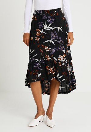 Kvinde iført en sort midi-nederdel med blomsterprint og flæsekant, kombineret med hvid langærmet top og hvide mulehælssko med spids tå.