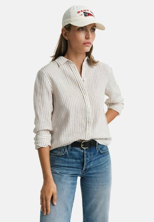 Camicia button-up a righe beige chiaro con colletto, abbinata a jeans in denim blu e un cappellino bianco con lettere rosse e un logo.