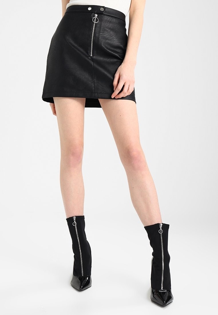 Femme portant une mini-jupe noire en similicuir avec fermeture éclair sur le devant et des bottines noires avec fermeture éclair devant sur un fond clair uni.