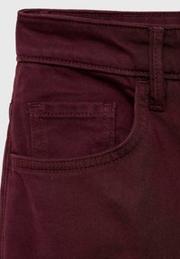 Tissu en denim bordeaux avec une finition texturée, comportant une poche avant et des passants de ceinture. Les coutures sont assorties en couleur.